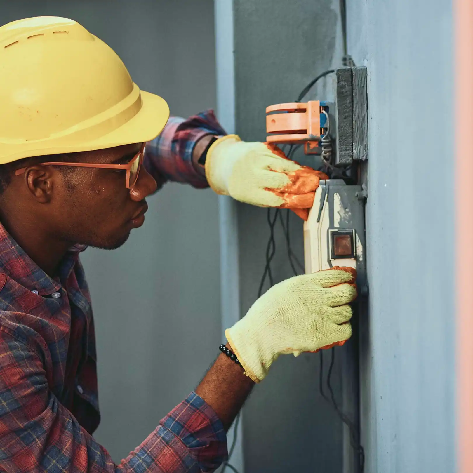 Homem consertando um painel elétrico, usando luvas de proteção e capacete de segurança em ambiente de trabalho.